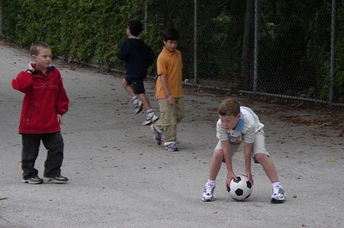 Boy in white shirt and shorts bending down to pick up soccer ball in schoolyard with another boy in a red jacket and another boy in an orange short walking up to him